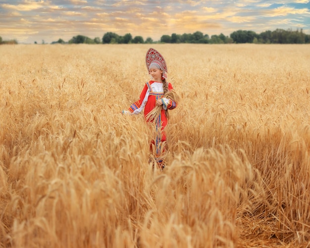 Kleines Mädchenkind im russischen nationalen Sarafan und ein Kokoshnik, der am Sommertag auf einem goldenen Weizenfeld steht