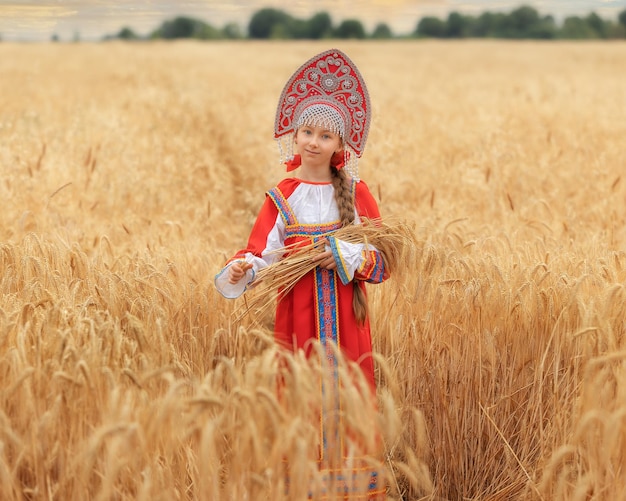 Kleines Mädchenkind im russischen nationalen Sarafan und ein Kokoshnik, der im Sommer auf einem goldenen Weizenfeld steht