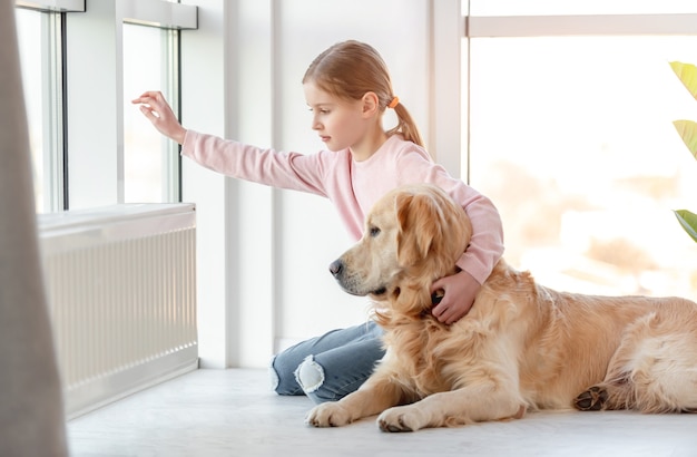 Kleines Mädchenkind mit goldenem Retrieverhund, der auf dem Boden sitzt und aus dem Fenster schaut