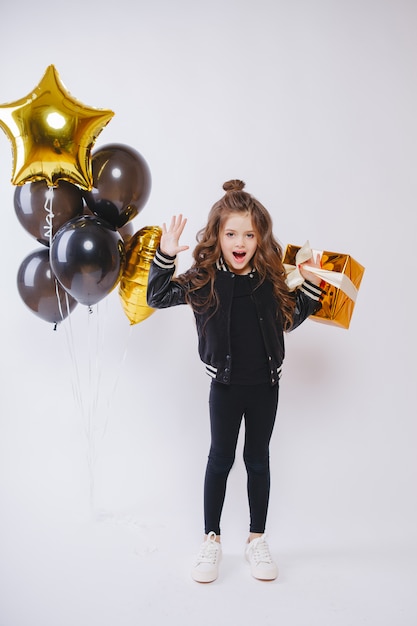 Kleines modernes Hipster-Mädchen in Modekleidung steht in der Nähe von Luftballons und hält Goldgeschenk. Pose Gesicht. Geburtstag.
