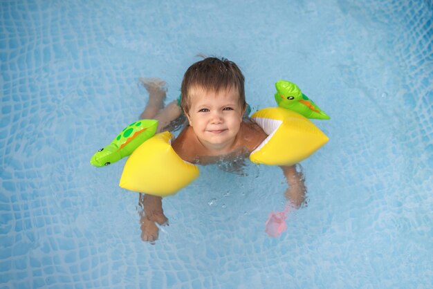 Kleines nacktes lächelndes Kind in grünen Badehosen mit hellen aufblasbaren Sicherheitsüberärmeln schwimmt und springt im Pool, im Hof, in transparentes, sauberes, kühles Wasser