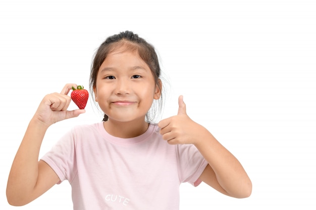Kleines nettes asiatisches Mädchen, das eine Erdbeere hält und sich Daumen zeigt,