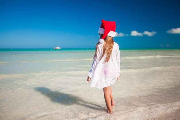 Kleines nettes Mädchen im roten Hut Weihnachtsmann auf dem Strand