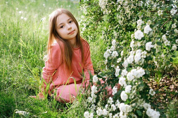 kleines nettes Mädchen in einem rosa Veloursanzug im Frühjahr im Park in der Nähe eines blühenden Busches mit Blumen