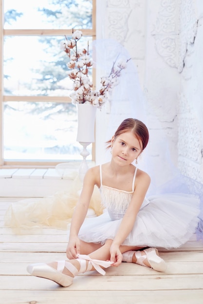 Kleines Primaballett. Junges Ballerinamädchen bereitet sich auf eine Ballettaufführung vor. Mädchen in einem weißen Ballkleid und Pointe am Fenster, schöne rote Haare. Junge Theaterschauspielerin