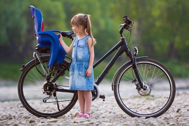 Kleines recht blondes Mädchen im blauen Kleid, das auf Kieseln vor modernem Fahrrad mit Kindersitz auf unscharfem grünem Baumhintergrund am Sommertag steht. Aktiver Lebensstil und Familienerholungskonzept.