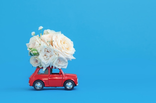 Kleines rotes Spielzeugauto mit Blumenstrauß Rosen auf dem Dach vor blauem Hintergrund Lieferung von Geschenken zum Valentinstag