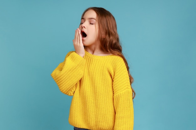 Kleines schläfriges Mädchen, das den Mund mit der Hand bedeckt, während es mit geschlossenen Augen gähnt, schläfriges Kind wacht auf und trägt einen gelben Pullover im Freizeitstil. Studioaufnahme im Innenbereich isoliert auf blauem Hintergrund.