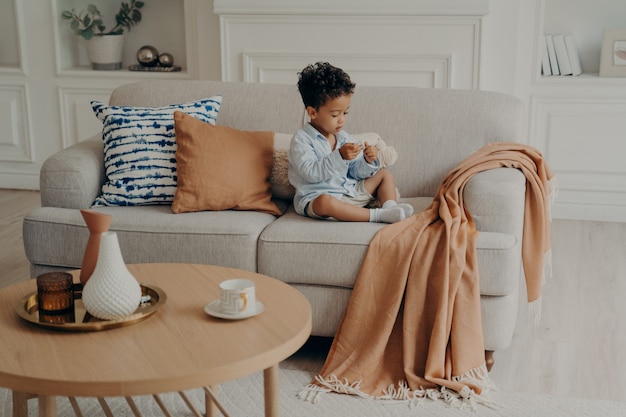 Kleines schönes afroamerikanisches Kind, das auf dem Sofa sitzt und alleine im Wohnzimmer spielt?