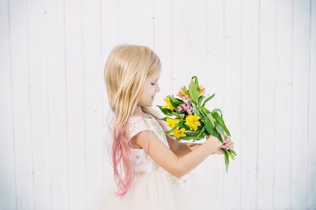 Kleines schönes Kindermädchen im schönen Kleid mit Blumenstrauß auf weißem hölzernem Hintergrund