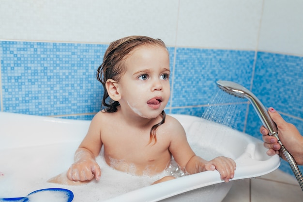 Kleines schönes Mädchen badet in einem Badezimmer, spielt den Narren und zeigt Zunge.