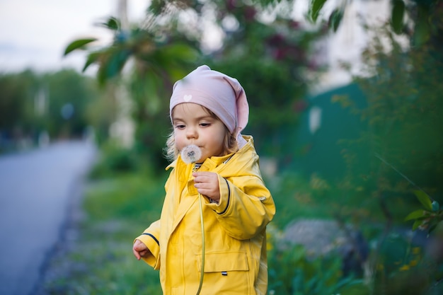 Kleines schönes Mädchen in einer rosa Mütze und einer gelben Jacke sitzt in einem Feld aus gelben Löwenzahn und Blumen