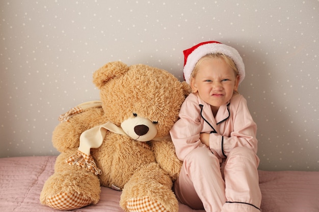 Kleines schönes Mädchen in rosa Pyjamas und Weihnachtsmütze sitzt gerne mit einem Teddybären auf dem Bett