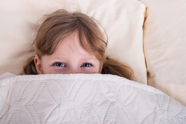 Kleines schönes Mädchen mit blauen Augen, die unter der Decke hervorschauen