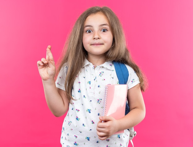 Kleines schönes Mädchen mit langen Haaren mit Rucksack, das ein Notizbuch hält, glücklich und überrascht, dass der Wunsch die Finger kreuzt, die auf Rosa stehen