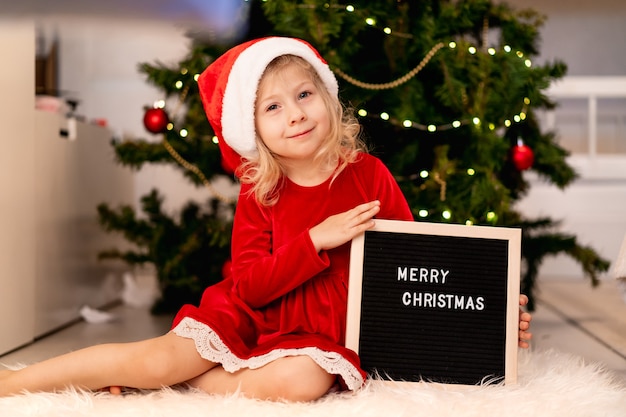 Kleines schönes Mädchen mit Weihnachtsmannmütze und rotem Weihnachtskleid liegt unter dem Weihnachtsbaum und lacht, neben einer schwarzen Tafel und weißen Buchstaben Text frohes neues Jahr 2021. Hochwertiges Foto