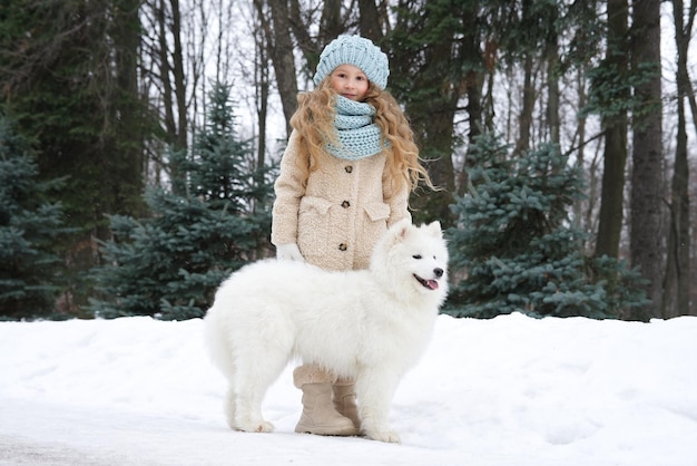 Kleines schönes Mädchen, süßes, glückliches, positives Kind, liegt auf dem Schnee und hat Spaß mit seinem großen weißen Hund an einem Wintertag im Park oder Wald