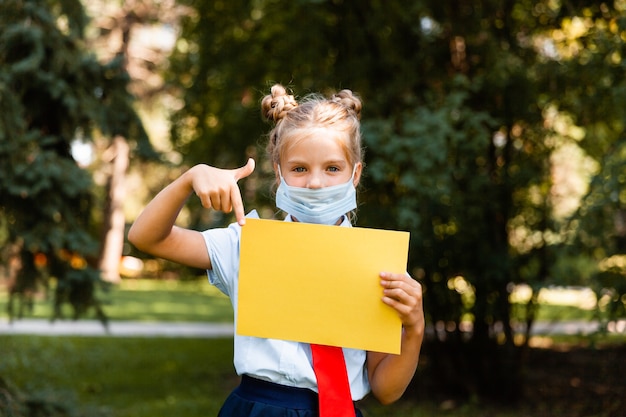 Kleines Schulmädchen in einer medizinischen Maske sitzt im Park auf dem Gras und hat Spaß.