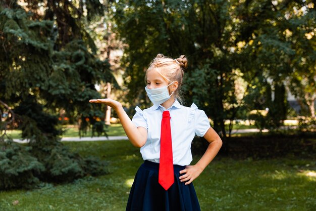 Kleines Schulmädchen in einer medizinischen Maske sitzt im Park auf dem Gras und hat Spaß.