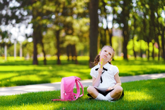 Kleines Schulmädchen mit dem rosa Rucksack, der auf Gras nach Lektionen und denkenden Ideen sitzt, las Buch- und Studienlektionen und schrieb Anmerkungen, Bildung und lernte Konzept