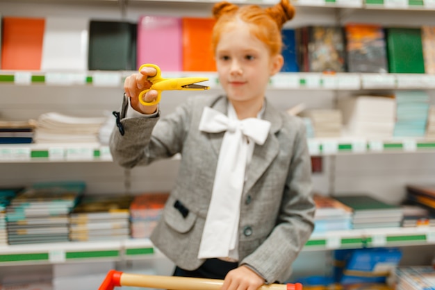 Kleines Schulmädchen mit einem Wagen, der eine Schere auswählt, im Schreibwarengeschäft einkaufen?