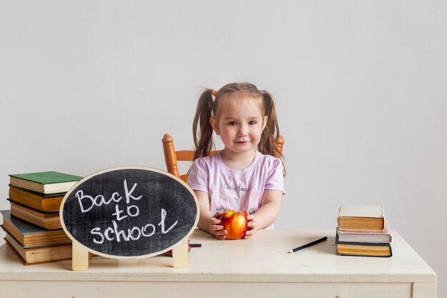 Kleines Schulmädchen sitzt an ihrem Schreibtisch in der Schule mit Lehrbüchern und hält Apfel