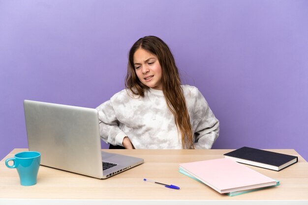 Kleines Studentenmädchen an einem Arbeitsplatz mit einem Laptop auf violettem Hintergrund, der an Rückenschmerzen leidet, weil er sich bemüht hat