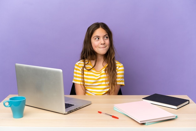 Kleines Studentenmädchen an einem Arbeitsplatz mit einem Laptop auf violettem Hintergrund, der beim Nachschlagen Zweifel hat