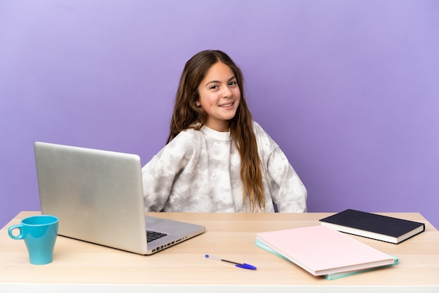 Kleines Studentenmädchen an einem Arbeitsplatz mit einem Laptop, der auf lila Wand isoliert ist, posiert mit Armen an der Hüfte und lächelt