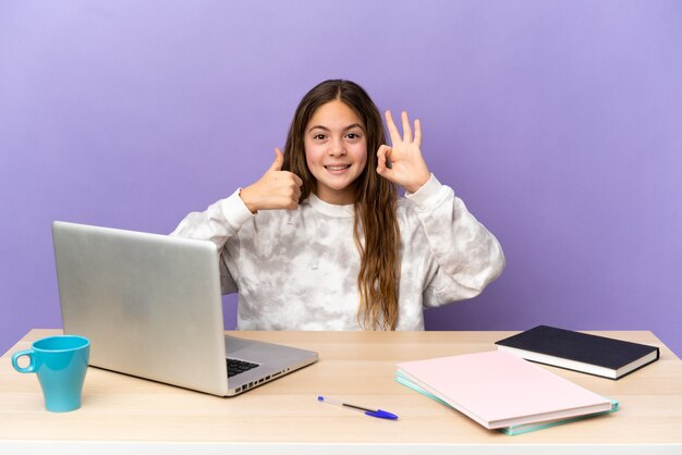 Kleines Studentenmädchen an einem Arbeitsplatz mit einem Laptop, der auf violettem Hintergrund isoliert ist und ein OK-Zeichen und eine Daumen-Hoch-Geste zeigt