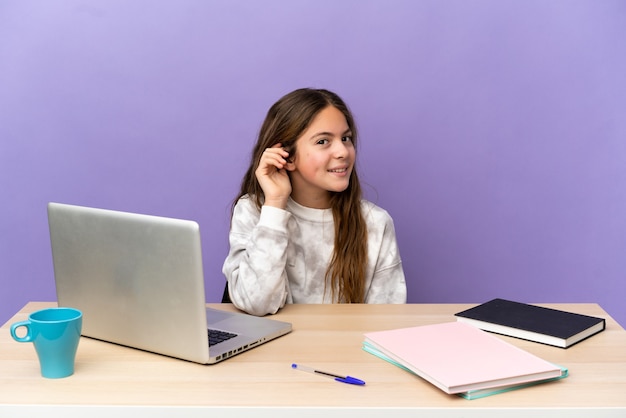 Kleines Studentenmädchen an einem Arbeitsplatz mit einem Laptop isoliert auf violettem Hintergrund, der etwas hört, indem er die Hand auf das Ohr legt