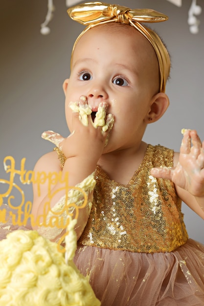 Kleines süßes Baby in einem goldenen Kleid mit einer Schleife auf dem Kopf, die einen jazzigen Geleekuchen von einer Creme versucht. Studioaufnahme eines Geburtstages auf einer grauen Wand, umgeben von Kugeln