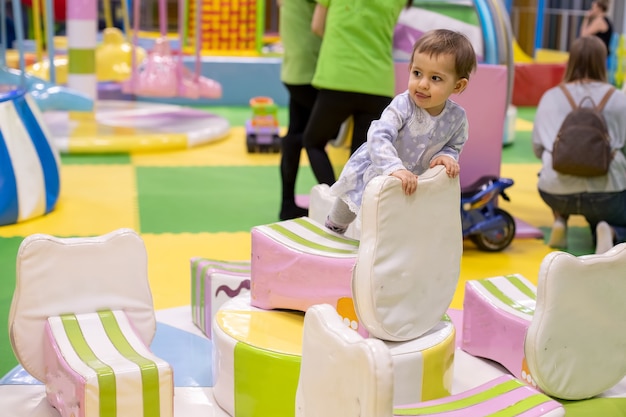 Kleines süßes Baby-Kleinkind-Mädchen in einem Kleid klettert auf Karussell im Kindertagesstätten-Spielzimmer