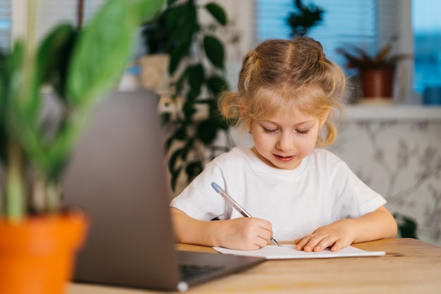 Kleines süßes blondes Mädchen schreibt, studiert zu Hause vor einem Laptop, Online-Lernen, zurück zur Schule.