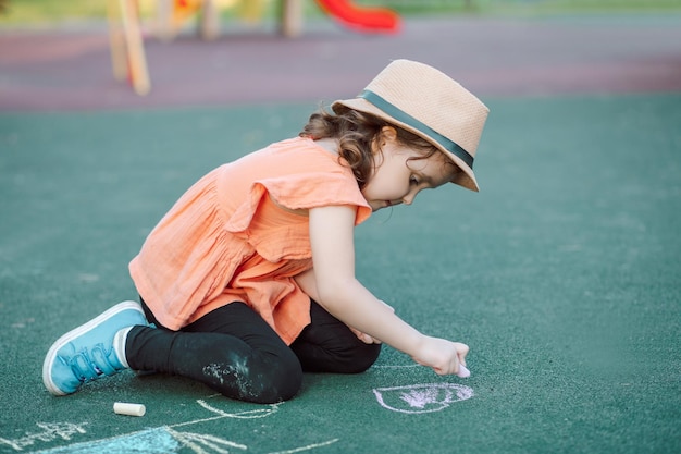 Kleines süßes glückliches Mädchen malt eine Kreide auf Asphalt auf dem Boden auf dem Spielplatz Kreative Sommeraktivität für Kinder