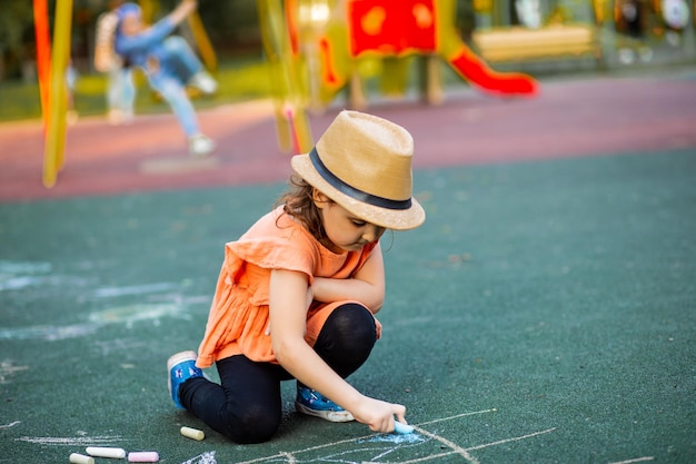 Kleines süßes glückliches Mädchen malt eine Kreide auf Asphalt auf dem Boden auf dem Spielplatz Kreative Sommeraktivität für Kinder