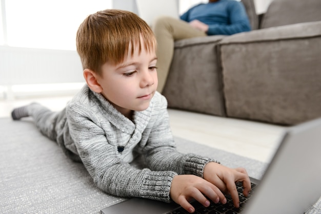 kleines süßes Kind mit Laptop, während es drinnen im Boden liegt. Schauen Sie sich den Laptop an.