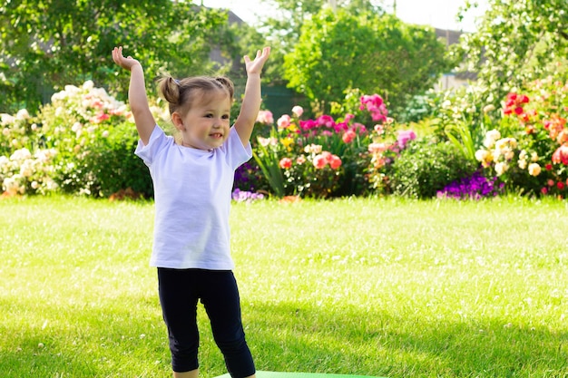 Kleines süßes lächelndes Mädchen 1-3 in einem weißen T-Shirt steht mit erhobenen Händen auf dem Gras vor dem Hintergrund von Blumen