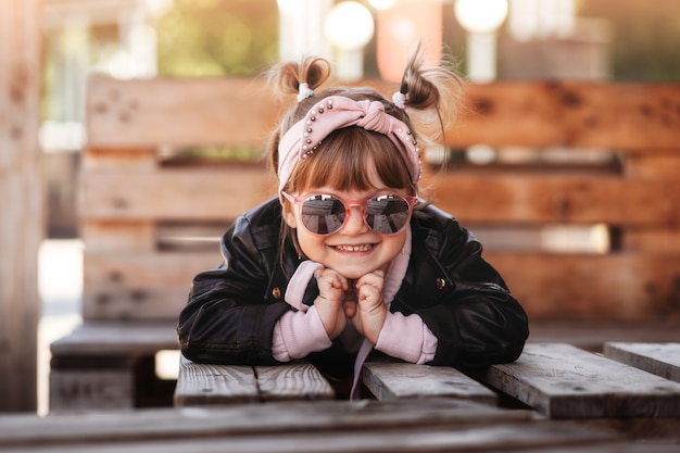 Kleines süßes lächelndes Mädchen mit Sonnenbrille sitzt auf der Terrasse des Cafés