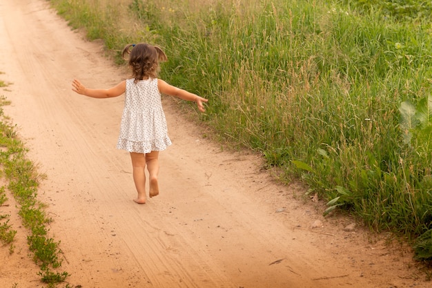 Kleines süßes Mädchen 1-3 in einem leichten Kleid, das im Sommer auf einem Weg auf dem Feld auf einem Grashintergrund läuft