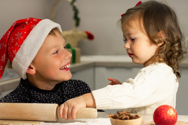 Kleines süßes Mädchen 2-4 mit roter Schleife und Junge 7-10 in einer Weihnachtsmütze machen Lebkuchen in der Neujahrsküche.