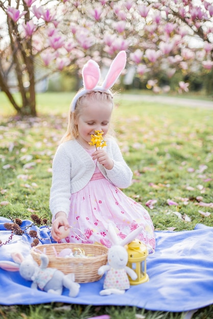 Kleines süßes Mädchen, das auf dem Gras nahe der Magnolie sitzt. Ein als Osterhase gekleidetes Mädchen hält eine Blume und ein Ei. Frühling.