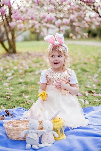 Kleines süßes Mädchen, das auf dem Gras nahe der Magnolie sitzt. Ein als Osterhase gekleidetes Mädchen hält eine Blume und ein Ei. Frühling.