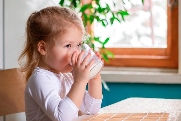 Kleines süßes Mädchen, das Milch trinkt, Kind, das morgens mit einem Glas heißer Milch frühstückt