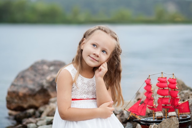 Kleines süßes Mädchen des Nahaufnahmeporträts im weißen Kleid und in den roten Segeln. Kind sitzen auf dem Seeweg mit Spielzeugschiff.