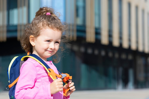 Foto kleines süßes mädchen geht in die erste klasse. glückliches kind zurück zur schule. kind mit rucksack, schultasche auf blauem hintergrund auf capmus draußen.