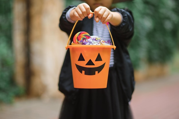 Kleines süßes Mädchen im Hexenkostüm mit Jackolantern-Kürbiseimer mit Bonbons und Süßigkeiten Kindertrick oder -behandlung im Halloween-Urlaub