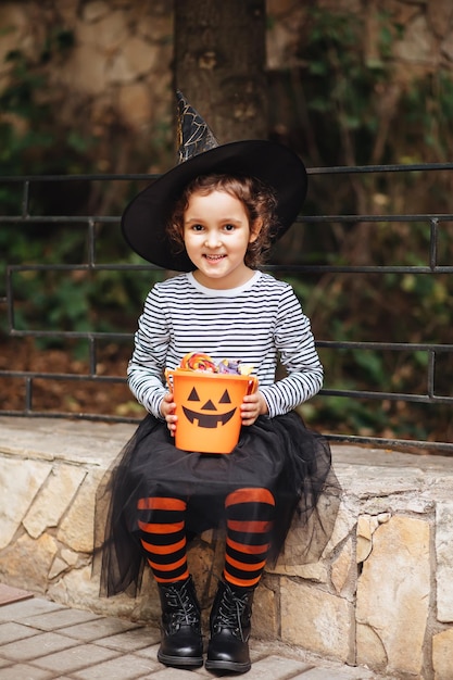 Kleines süßes Mädchen im Hexenkostüm mit Jackolantern-Kürbiseimer mit Bonbons und Süßigkeiten Kindertrick oder -behandlung im Halloween-Urlaub