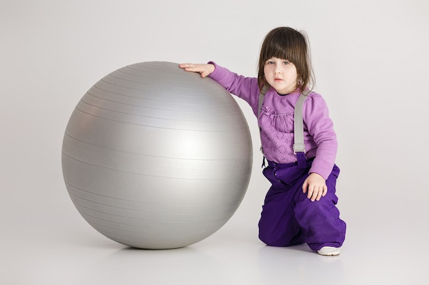 Kleines süßes Mädchen in den lila Kleidern mit einem großen Ball für Fitness auf grauem Hintergrund.