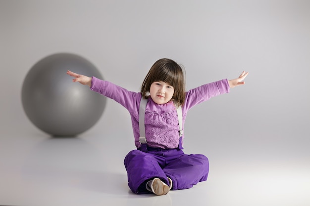 Kleines süßes Mädchen in lila Kleidern mit ausgestreckten Armen und mit einem großen Ball für Fitness auf grauem Hintergrund.